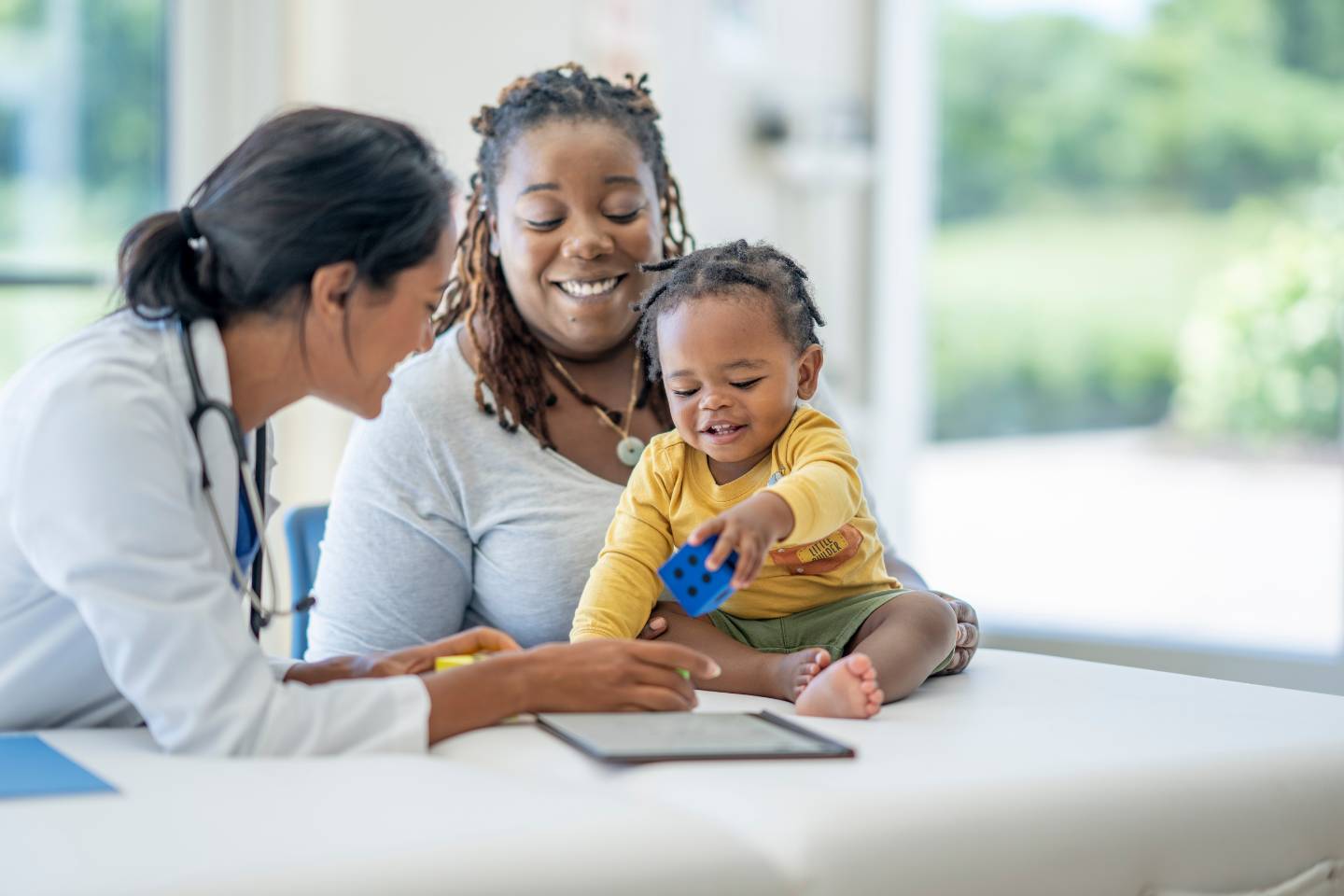 A doctor interacts with a baby holding a blue toy, sitting on a table with their mother, as the dynamic content of their joyful expressions creates an atmosphere of warmth and trust.