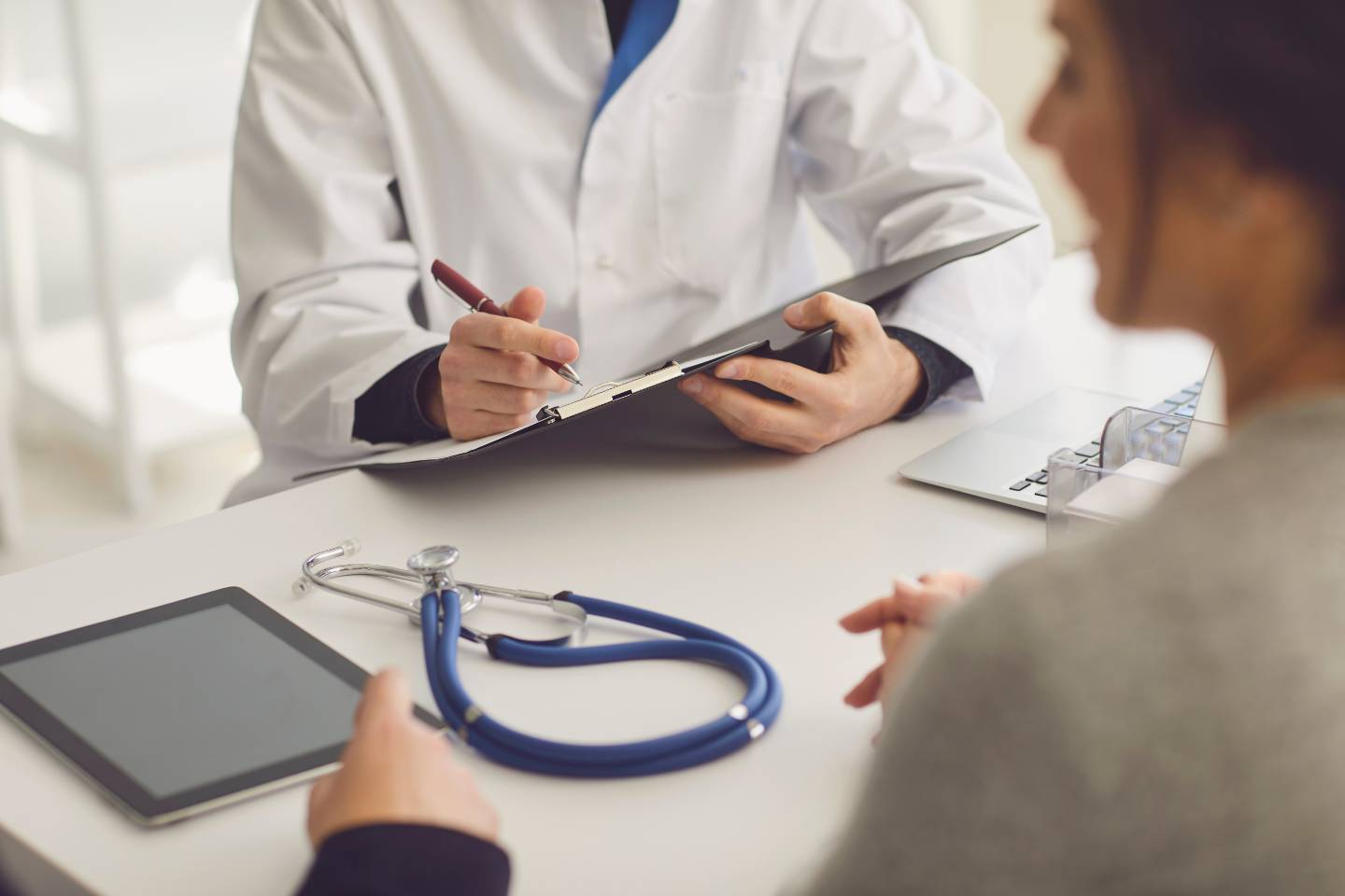A doctor in a white coat writes on a clipboard while discussing dynamic content with a patient. A stethoscope and tablet lie on the desk.