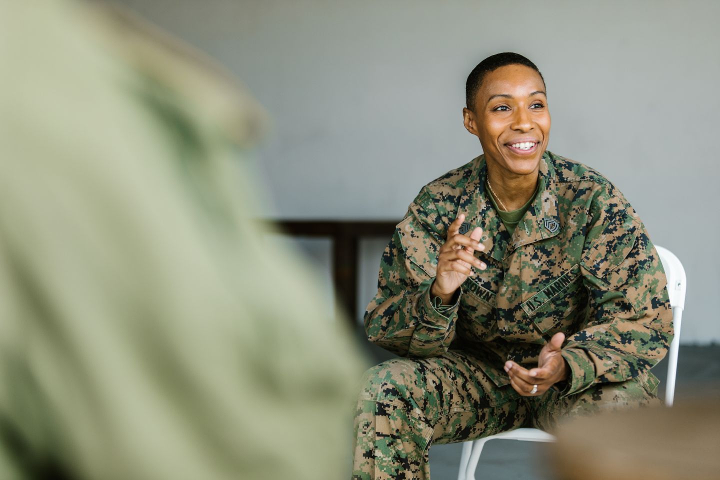 In a room filled with dynamic content, a person in military uniform sits and gestures while smiling, their demeanor as engaging as the surroundings.